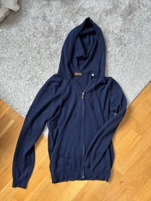 Merino zip från Stenströms - Säljer en mörkblå hoodie från Stenströms med dragkedja och huva. Tröjan är tillverkad i ett mjukt stickat material och har ribbade muddar vid ärmslut och nederkant. Perfekt för en avslappnad och stilren look.