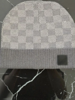 Grå rutig mössa med patch - Snygg grå mössa med rutigt mönster och ribbad kant. Mössan har en diskret svart patch framtill och är tillverkad i ett mjukt stickat material. Perfekt accessoar för kalla dagar och passar till de flesta outfits.