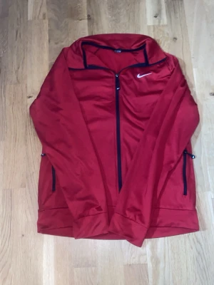 Röd träningsjacka från Nike - Röd träningsjacka från Nike med hel dragkedja och svarta detaljer. Jackan har hög krage, långa ärmar och två sidofickor med dragkedja. Perfekt för löpning eller gym. Materialet är lätt och andas, vilket gör den bekväm under träning.