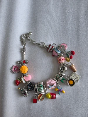 Färgglatt berlockarmband med charms - Unikt armband i silverfärgad metall med massor av färgglada berlocker i form av t.ex. tårta, körsbär, kamera, blommor och kaffe. Armbandet har detaljer i rosa, gult, rött, grönt och blått. Perfekt för dig som gillar personliga och lekfulla accessoarer.