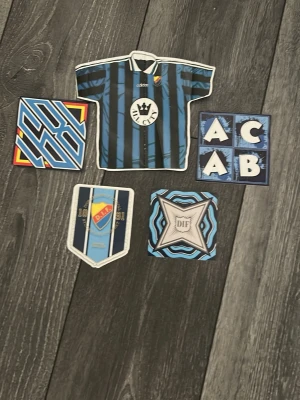 Djurgården klistermärken samling - Säljer en samling klistermärken med Djurgården-tema. Motiven inkluderar en tröja, DIF-logga, blåa och svarta mönster samt text. Perfekt för att dekorera laptop, mobil eller andra ytor. Färgerna är främst blå, svart, vit och lite röd och gul.