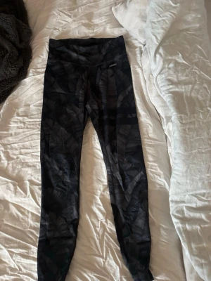 Svarta tights med mönster från Stronger - Snygga svarta träningstights från Röhnisch med diskret bladliknande mönster. Hög midja och stretchigt material som passar perfekt för gym, löpning eller yoga. Tightsen har en liten logga framtill och en mobilficka baktill