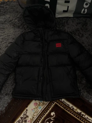 Svart dunjacka med huva från Hugo - Säljer en svart dunjacka från Hugo med huva och röd logotyp på bröstet. Jackan har quiltad design, dragkedja och knappar framtill. Perfekt för kalla vinterdagar och riktigt snygg att bära till jeans eller joggers. Jackan är nästan helt ny, har haft på mig den 3 gånger. Bilderna där jag har haft på mig den finns med på annonsen. Jag säljer jackan eftersom den är för liten för mig. 