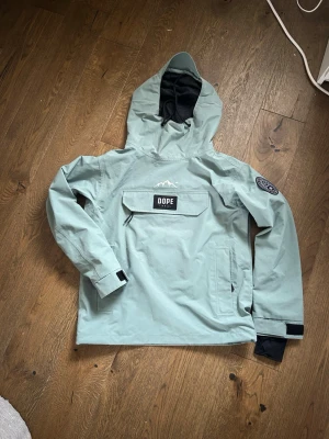 Ljusgrön vindjacka från Dope Snow - Snygg ljusgrön vindjacka från Dope Snow med stor magficka, huva och svart patch på ärmen. Jackan har dragkedja vid sidan, kardborre vid ärmslut och svart innerfoder i huvan. Perfekt för dig som gillar streetwear och outdoor.