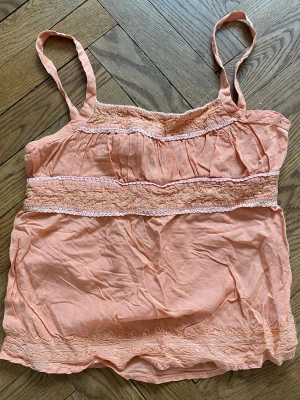 Persikofärgad broderad linnetopp - Så fin persikofärgad linnetopp med tunna axelband och broderade, glittriga detaljer i matchande ton. Toppen har en avslappnad passform och dekorativa band under bysten och vid fållen samt knytning baktill. Perfekt för varma dagar och enkel att styla med jeans eller kjol🧡🌷
