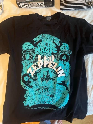 Led Zeppelin svart t-shirt - Säljer en svart t-shirt med stort turkost och vitt Led Zeppelin-tryck på framsidan. T-shirten har klassisk rund hals och korta ärmar. Perfekt för dig som gillar bandtröjor och vill sticka ut med en cool retrodesign.