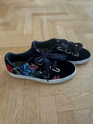 Svarta sneakers med blommig brodyr Puma - Unika svarta sneakers från Puma i sammet med stora satinband som snörning och färgglad blommig brodyr på sidan. Skorna har vita sulor och guldfärgade öljetter som ger en extra touch. 