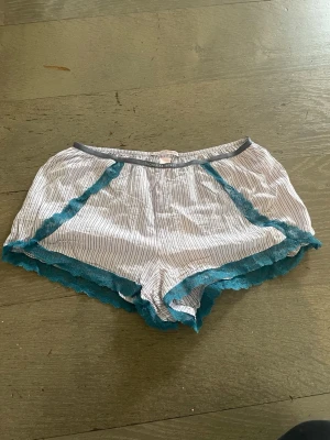 Randiga pyjamasshorts med spets - Säljer ett par vita pyjamasshorts från Victoria's Secret med tunna svarta ränder och turkos spetsdetalj längs kanterna. Shortsen har en avslappnad passform och elastisk midja för extra komfort. Perfekta för mysiga hemmakvällar.