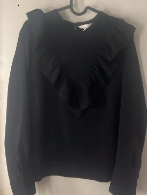 Svart blus med volangdetalj - Snygg svart blus med lång ärm och volang i V-form framtill. Blusen har rund halsringning och är tillverkad i ett lätt tyg som ger ett stilrent intryck. Perfekt för dig som gillar trendiga detaljer och enkel design.