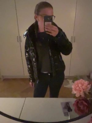 Svart glansig pufferjacka med dragkedja - Säljer en svart pufferjacka med glansig finish och två framfickor med dragkedja. Jackan har hög krage och quiltad design, perfekt för kalla dagar. Materialet är syntet och jackan har en cool, modern look som passar till jeans och sneakers.