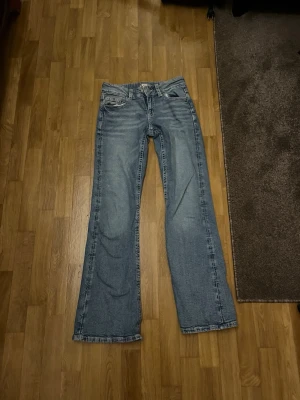 Ljusblå bootcut jeans med broderi - Säljer ett par ljusblå bootcut jeans med snygga broderade detaljer på bakfickorna och knappar. Klassisk femficksmodell med låg midja och slitna detaljer. Perfekta för dig som gillar en avslappnad och trendig stil.