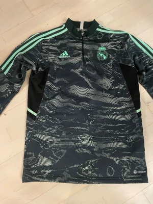 Real Madrid träningströja Adidas - Snygg långärmad träningströja från Adidas med Real Madrid-logga. Svart bas med mintgröna detaljer och mönster, halv dragkedja och klassiska Adidas-ränder på ärmarna. Tillverkad i funktionsmaterial som andas, perfekt för träning.