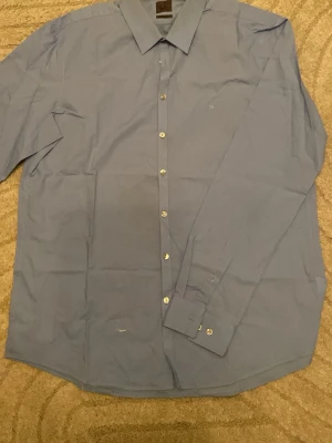 Blå skjorta från Calvin Klein - Snygg blå skjorta från Calvin Klein med klassisk krage och diskret 'ck'-brodyr på bröstet. Skjortan har långa ärmar, knappar framtill och är i en slim passform. Perfekt för dig som gillar stilrena och enkla plagg.