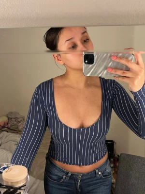 Mörkblå croppad randig topp - Säljer en mörkblå croppad topp med vita vertikala ränder och djup v-ringning. Toppen har lång ärm och är figurnära i modellen. Perfekt för dig som gillar trendiga och bekväma plagg med lite edge.