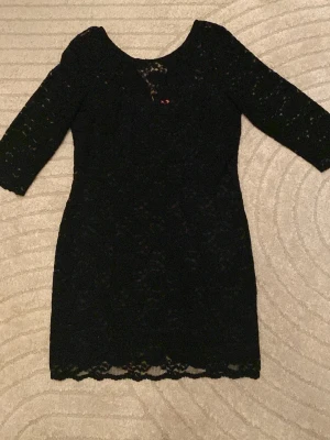 Svart spetsklänning med 3/4-ärm - Snygg svart klänning i spets med 3/4-ärm och rund halsringning. Klänningen har en guldfärgad dragkedja i ryggen och fina spetsdetaljer längs nederkanten. Figurnära passform och helfodrad för en elegant look.