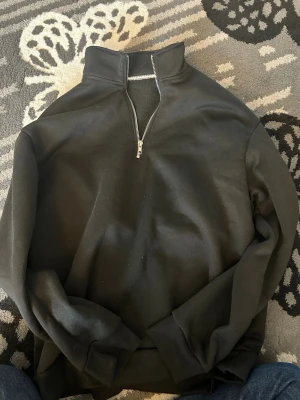 Svart halvzip hoodie - Snygg svart hoodie med halv dragkedja och hög krage. Tröjan har ribbade muddar vid ärmslut och nederkant. Perfekt för en avslappnad och stilren look. Materialet känns mjukt och bekvämt mot huden.
