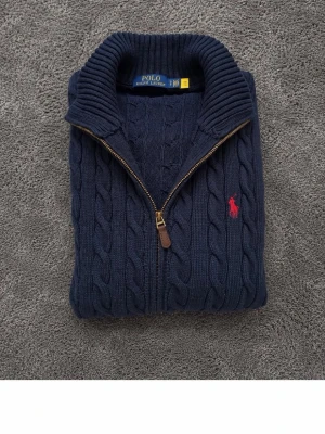Mörkblå kabelstickad kofta Polo Ralph Lauren - Mörkblå kabelstickad kofta från Polo Ralph Lauren med dragkedja och hög krage. Klassisk röd broderad logga på bröstet och ribbade muddar. Perfekt för lager-på-lager och snygg till jeans. Materialet är mjukt och håller värmen. Medföljer tags. Har ingen stress med att sälja så inga skambud.