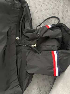 Svart vindjacka från Moncler - Snygg svart vindjacka från Moncler med dragkedja framtill och huva med dragsko. Ärmarna har röd, vit och blå randdetalj. Jackan är tillverkad i ett lätt, vattenavvisande material och har Moncler-logga på ena ärmen. Perfekt för blåsiga dagar. Liten skada på dragskedjan M som passar som S