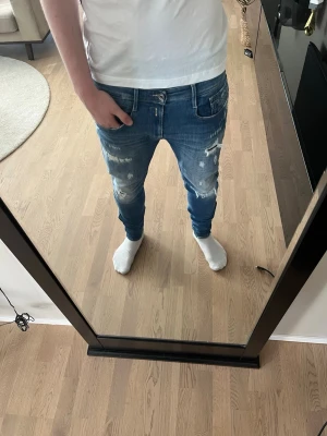 Replay Jeans  - Säljer ett par blå replay jeans med slitningar. Jeansen har klassisk femficksdesign, dragkedjegylf och är i stretchigt denimtyg. Perfekta för en avslappnad och trendig look.