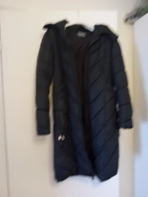 Svart lång dunjacka med huva - Snygg svart lång dunjacka med huva och dragkedja framtill. Jackan har ett quiltat mönster i diagonala linjer och är perfekt för kalla vinterdagar. Praktiska fickor på sidorna och en stilren look som passar till det mesta.