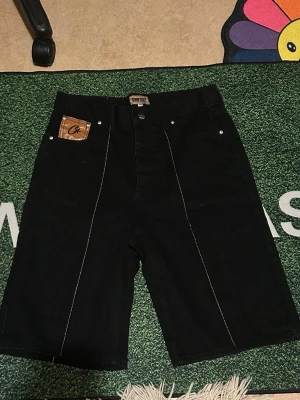 Svarta shorts från Corteiz - Svarta shorts med vita kontrastsömmar och broderade stjärndetaljer på bakfickorna. Klassisk femficksmodell med läderpatch framtill och baktill. Raka ben och midwaist passform, perfekta för en streetwear-look.