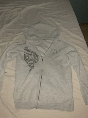 Grå zip-hoodie med paljettdetaljer - Snygg ljusgrå hoodie från Bvaldi med dragkedja och huva. Framsidan har glittriga paljettdetaljer och logga på bröstet. Perfekt för en chill och trendig look. Mjuk bomullsblandning och långa ärmar. Nypris 1200kr 