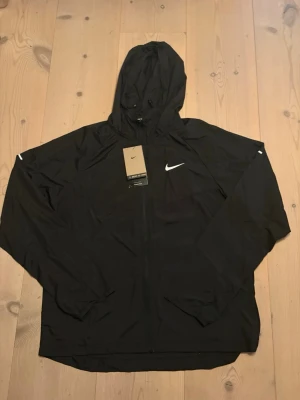 Svart vindjacka från Nike med huva - Svart vindjacka från Nike med vit logga på bröstet och huva. Jackan har dragkedja framtill, långa ärmar och elastiska muddar. Tillverkad i lätt och tåligt material, perfekt för blåsiga dagar. Enkel och stilren design som passar till det mesta.