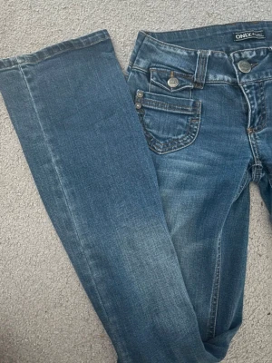 Blå jeans från ONLY med fickdetaljer - Snygga blå bootcut jeans från Only med  fickdetaljer framtill. Jeansen är lågmidjade och har lite stretch i sig. De har silverfärgade knappar och är gjorda i denim med lätt tvättad look. Perfekta för en avslappnad och trendig stil. Storlek 27/34!!