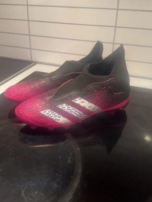 Adidas Predator fotbollsskor svart/röd - Säljer ett par Adidas Predator fotbollsskor i svart och röd med silvriga detaljer och mönstrad ovandel. Skorna har en strumpliknande passform och är tillverkade i syntetmaterial, perfekta för dig som vill sticka ut på planen.