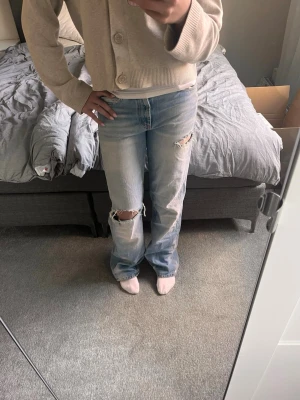 Ljusblå slitna raka jeans - Säljer ett par ljusblå jeans med raka ben och slitningar vid knäna och låret. Jeansen har medelhög midja. Perfekta för trendig look. Materialet är denim i bomull.