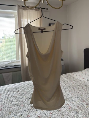 Beige linnetopp med drapering -  beige linnetopp med djup draperad urringning framtill. Toppen är ärmlös och har en mjuk, följsam passform som känns både trendig och stilren. Perfekt att matcha med jeans eller kjol för en cool look. 