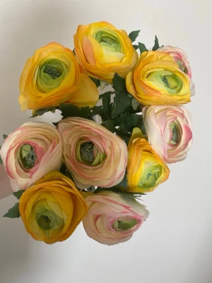 Bukett konstgjorda ranunkler i tyg  - Säljer en bukett med konstgjorda ranunkler i gult, vitt och rosa från Flowers by SIA. Blommorna har gröna blad och långa stjälkar i plast, perfekta som dekoration i hemmet. Realistiskt utseende och fina färgskiftningar.