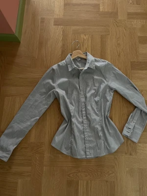  H&M skjorta  - Snygg skjorta med smala svarta och vita ränder, klassisk krage och knäppning framtill. Skjortan har figurnära passform och långa ärmar med manschett. Perfekt till både jeans och kostymbyxor.