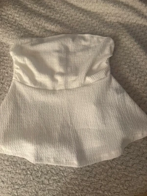 Vit bandeau tubtopp i strukturerat tyg - Säljer en vit bandeau tubtopp med lätt strukturerat tyg och utsvängd nederdel. Toppen är axelbandslös och har en stilren, enkel design som passar perfekt till jeans eller kjol.