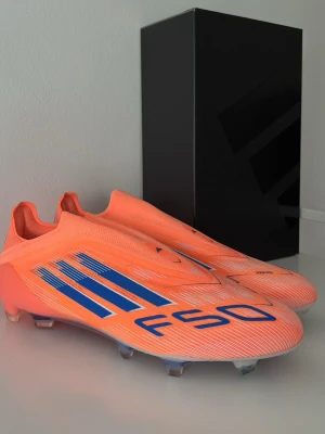 Adidas F50 CORAL BLAZE Laceless Elite FG - Snygga Adidas F50 fotbollsskor i en fräsch orange färg med blå detaljer och klassiska tre ränder på sidan. Skorna har låg profil, strömlinjeformad design och är tillverkade i syntetmaterial för bästa prestanda på planen. Perfekta för dig som vill sticka ut på matchen.