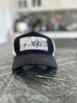 Svart lozama keps - Snygg lozama keps i svart och vitt med ett unikt svartvitt tryck framtill. Kepsen har böjd skärm och mesh på sidorna för extra ventilation. Perfekt accessoar för en avslappnad streetstyle-look.
