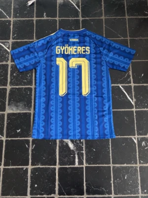 Sveriges Bortaställ VM 2026 | Gyökeres #17 - Säljer en blå Sverige fotbollströja från Adidas med gult tryck, nummer 17 och namnet Gyökeres på ryggen. Tröjan har korta ärmar, gul krage och tre gula ränder på axlarna samt Sveriges landslagsemblem på bröstet. Materialet är lätt och andas.