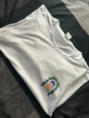 Vit Casablanca t-shirt med tryck - Vit t-shirt från Casablanca med färgglatt tryck av en byggnad och gröna blad på ryggen samt mindre tryck på bröstet. Rund halsringning och klassisk passform. Perfekt för dig som gillar stilrena plagg med unik design.
