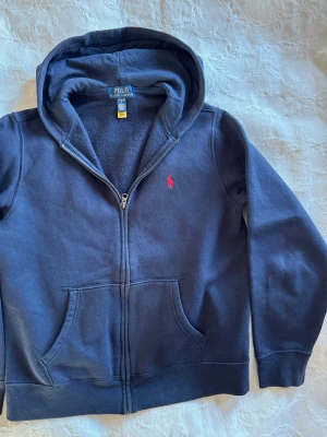 Mörkblå hoodie från Ralph Lauren - Mörkblå hoodie med dragkedja från Polo Ralph Lauren. Klassisk design med huva, kängurufickor och röd broderad logga på bröstet. Mjuk insida och ribbade muddar. Perfekt för en avslappnad och stilren look. Tröjan är i nyskick då den var för liten när sonen fick den.