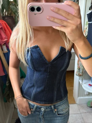 Mörkblå denim korsett-topp - Snygg mörkblå korsett-topp i denim med markerade sömmar och en unik v-ringad skärning framtill. Toppen är ärmlös och har en tight passform som framhäver midjan. Perfekt att styla med jeans eller kjol för en trendig look.