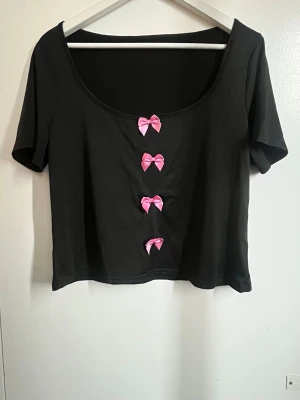 Svart croppad topp med rosa rosetter - Svart croppad topp med korta ärmar och fyra söta rosa rosetter framtill. Toppen har en rundad, djup urringning och är perfekt för en trendig och lekfull stil. Mjuk och stretchig känsla.