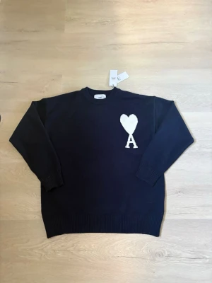 Mörkblå stickad tröja från Ami - Mörkblå stickad tröja från Ami med rund halsringning och ribbade muddar. På bröstet finns ett stort vitt hjärta och bokstaven 'A' som är typiskt för märket. Perfekt för dig som gillar stilrena och trendiga plagg. Priset här inte hugget i sten så priset kan diskuteras.