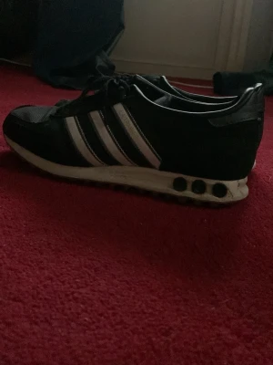 Svarta Adidas sneakers med vita ränder - Svarta Adidas la trainers med klassiska tre vita ränder på sidorna och vit sula med svarta detaljer. Ovandelen är i mesh och mockaliknande material, med svarta snören. I bra skick