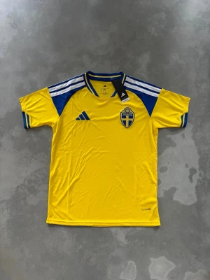 Sveriges hemmaställ 2026 - Gul fotbollströja med blå detaljer och Sveriges landslagsemblem på bröstet. Tröjan har korta ärmar, rund hals och klassiska Adidas-ränder på axlarna. Perfekt för dig som vill visa ditt stöd för Sverige på matchen eller träningen.