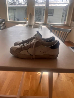 Vita sneakers från Saint Laurent - Säljer ett par klassiska vita sneakers från phillip model med marin blåa detaljer på hälen. Skorna har lågt skaft, vita skosnören och är tillverkade i läder med mockadetaljer. Perfekta för dig som gillar en stilren och tidlös look. PRISET ÄR EJ HUGET I STEN VID SNABB AFFÄR!