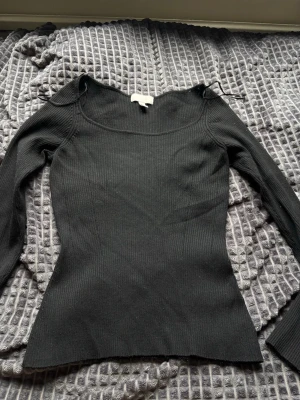 Svart ribbstickad topp med lång ärm - Säljer en svart, ribbstickad topp med lång ärm och bred, rundad urringning. Tröjan har en figurnära passform och är tillverkad i ett mjukt, stretchigt material. Perfekt att styla med jeans eller kjol för en clean look.