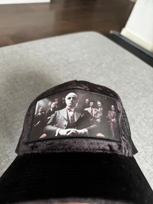 Svart keps med tryck av gruppfoto - Säljer en svart keps i sammet med mesh på sidorna och ett tryck framtill som föreställer ett gruppfoto av män i kostym. Kepsen har böjd skärm och justerbar baksida. Perfekt för dig som gillar unika accessoarer med coolt motiv.
