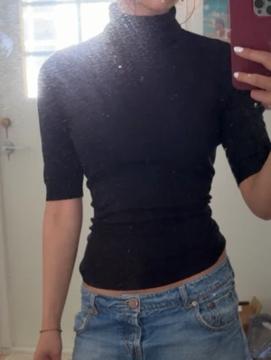 Svart polotopp med halvlånga ärmar - Snygg svart polotopp med tight passform och halvlånga ärmar. Toppen har hög krage och är tillverkad i ett stretchigt material som sitter skönt mot kroppen. Perfekt att styla med jeans eller kjol för en trendig look.