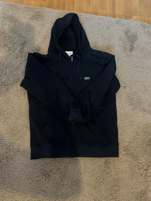 Lacoste Mörkblå ziptröja  - Säljer en mörkblå oanvänt zip hoodie från Lacoste med dragkedja och klassisk krokodillogo på bröstet som jag säljer pga att den blev för liten. Den är i storleken M men passar även i storleken S. Tröjan har huva med snören, ribbade muddar och två fickor framtill. Perfekt för en avslappnad och cool look.