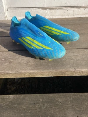 Adidas X Speedflow blå fotbollsskor - Säljer ett par Adidas X Speedflow fotbollsskor i blått med gula detaljer och klassiska tre ränder på sidorna. Skorna har låg vikt, strumpliknande passform och fasta dobbar för spel på gräs. Perfekta för dig som vill ha snabbhet och kontroll på planen. De är inte använda många gånger nästan som nya faktiskt.